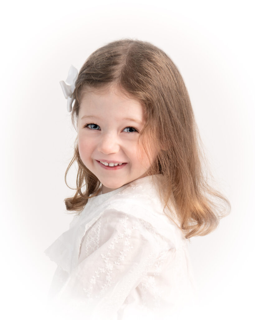 three year old girl in an heirloom portrait looking over her shoulder at the camera and smiling with a bow in her hair