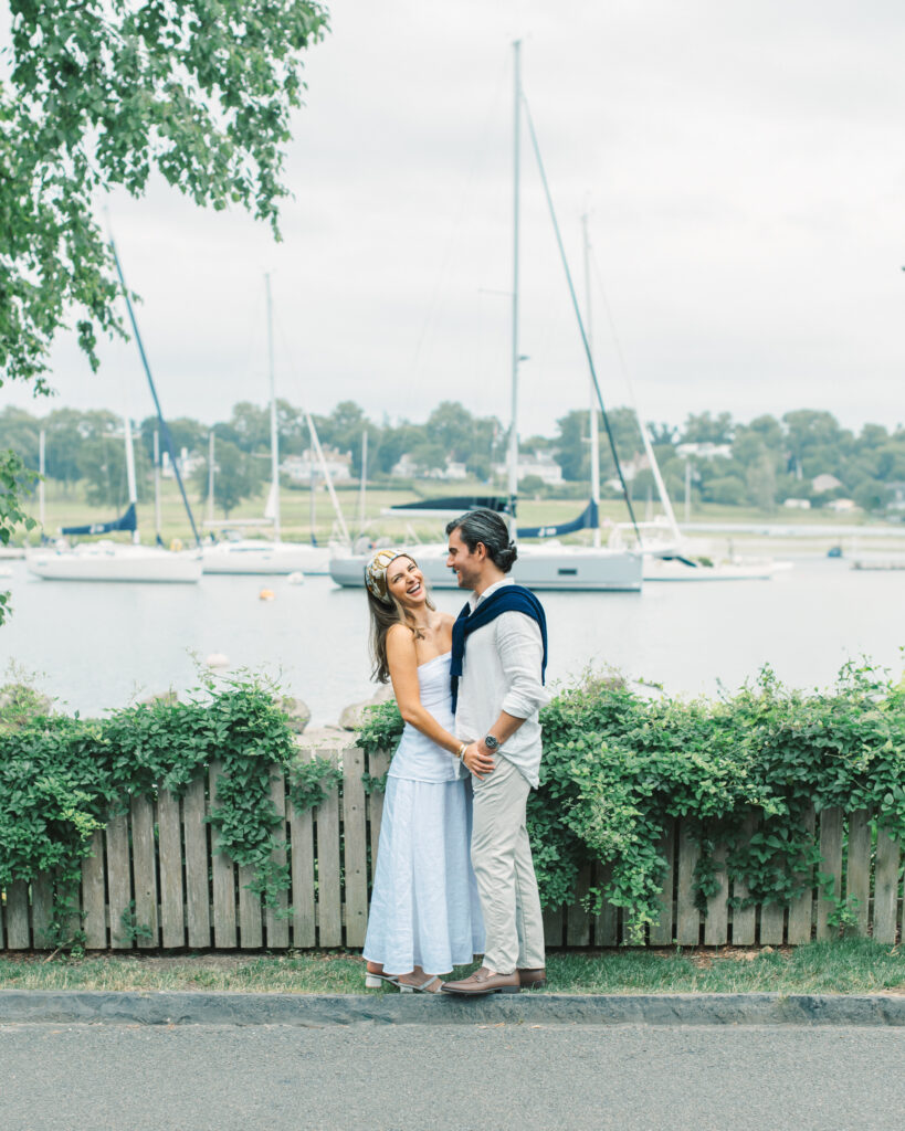 engaged couple smiling and laughing together in front of a beautiful sound with lots of sailboats in the background