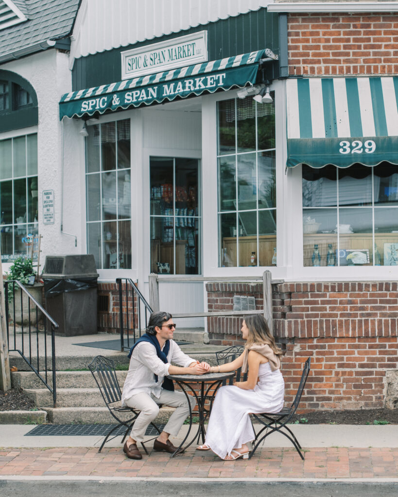 an engaged couple sitting at a table and holding hands in front of Spic & Span Market in Southport, CT